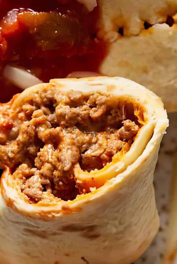 A close-up of a burrito filled with seasoned ground beef, topped with red salsa, being cut by a fork. Shredded cheese and herbs are visible in the background.