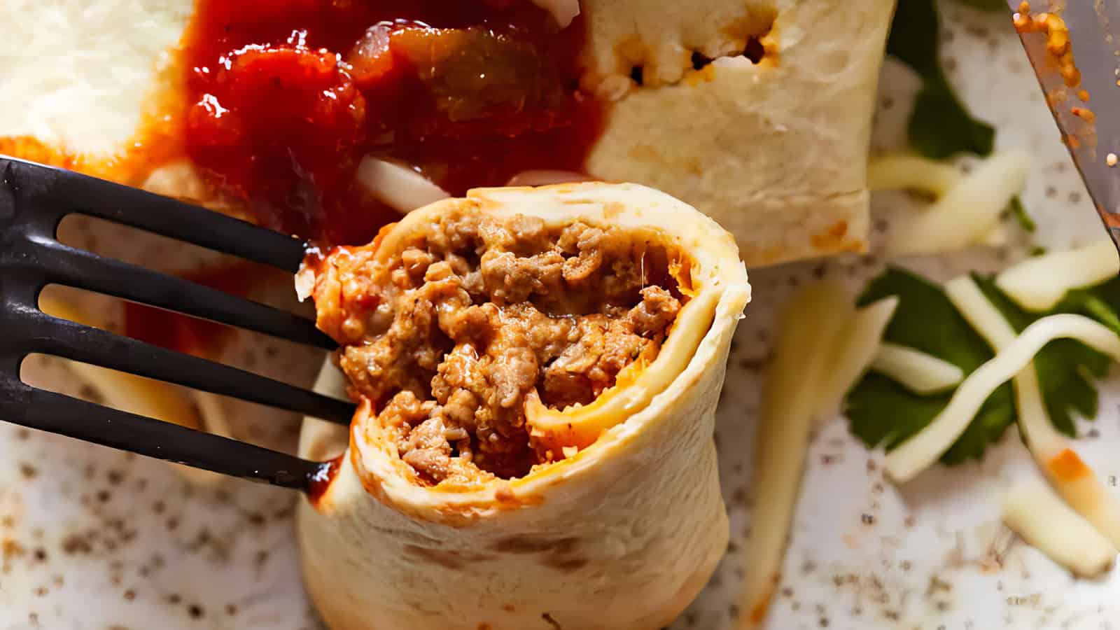 A close-up of a burrito filled with seasoned ground beef, topped with red salsa, being cut by a fork. Shredded cheese and herbs are visible in the background.