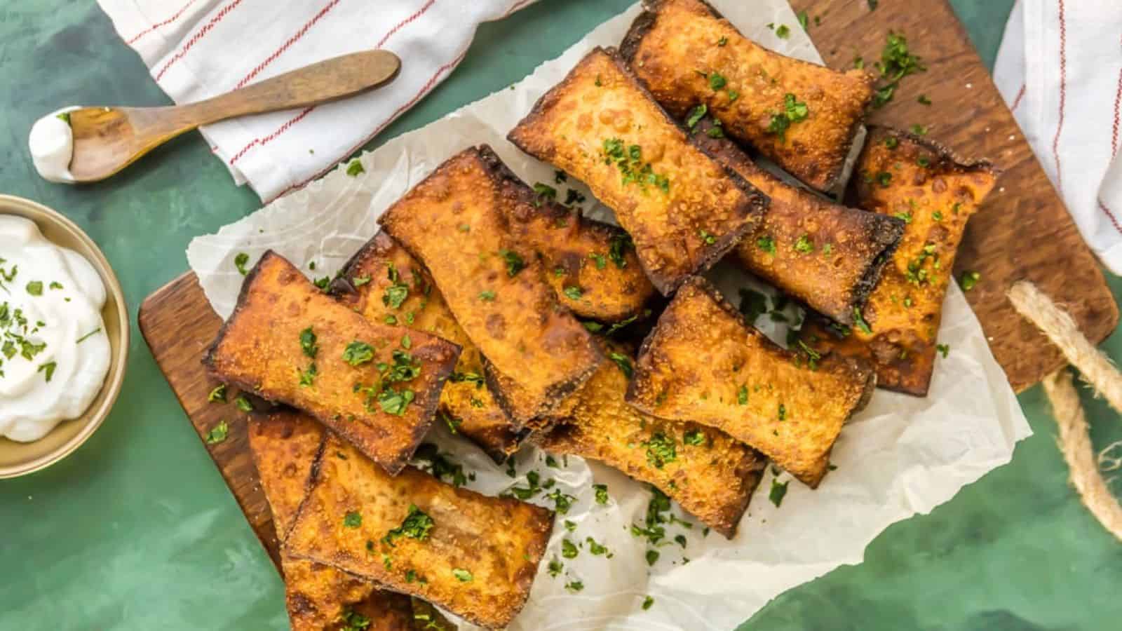A pile of crispy, golden-brown square-shaped snacks garnished with chopped herbs on a parchment-lined wooden board. A small bowl of white dip with herbs next to a spoon on a cloth.