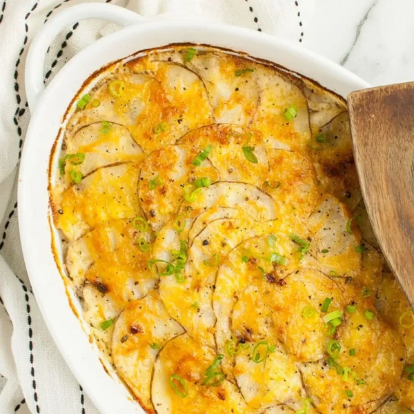 A white ceramic dish filled with baked scalloped potatoes topped with melted cheese and chopped green onions. A wooden spatula rests on the right side of the dish. The dish is placed on a white cloth with black stitching on a marble surface.