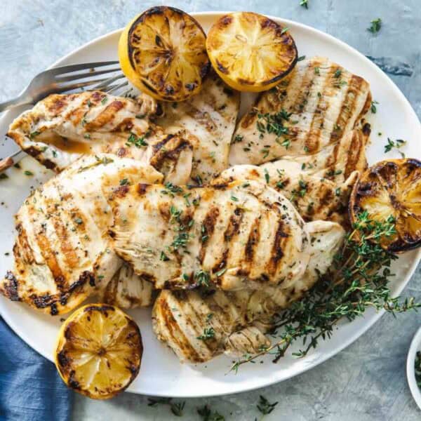 A white plate with grilled chicken breasts, charred lemon halves, and fresh herbs on a light gray surface.