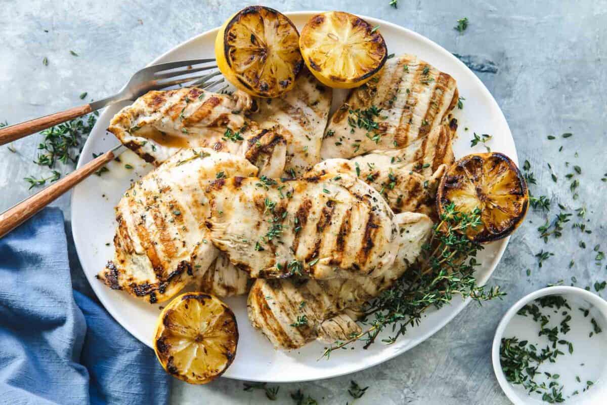 A white plate with grilled chicken breasts, charred lemon halves, and fresh herbs on a light gray surface.