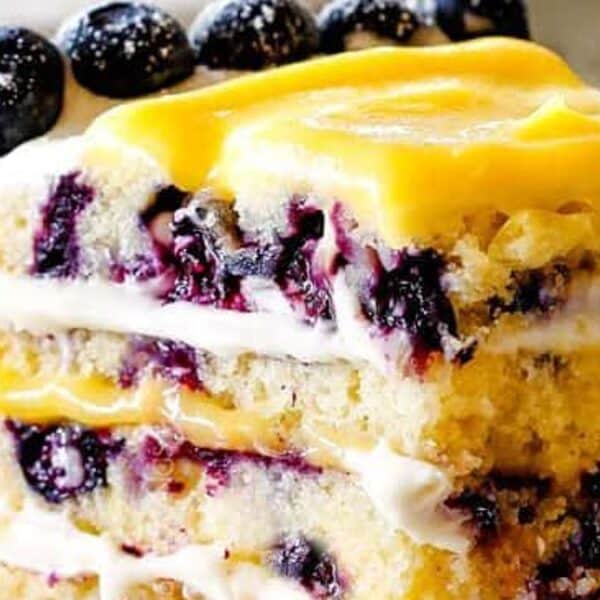A slice of layered cake with white frosting, yellow cream topping, and whole blueberries on top; additional blueberries are placed beside the slice.