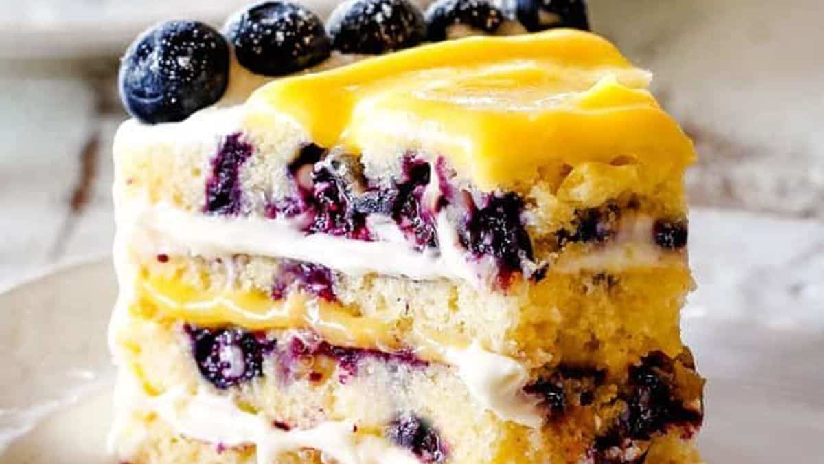 A slice of layered cake with white frosting, yellow cream topping, and whole blueberries on top; additional blueberries are placed beside the slice.