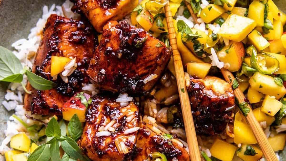 Grilled glazed chicken pieces on white rice, topped with fresh herbs and accompanied by a colorful mango salsa; chopsticks placed to the side.