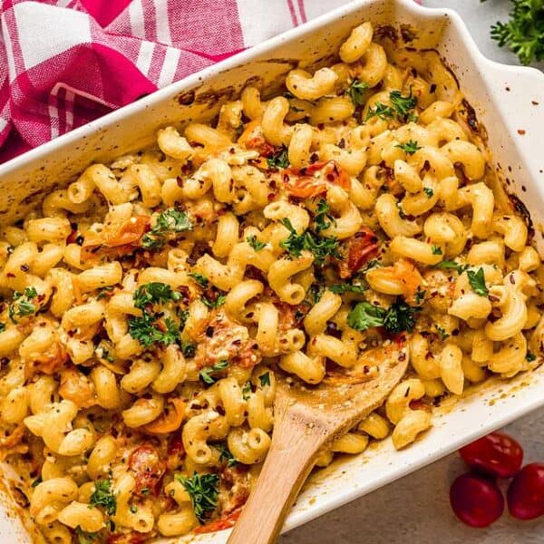 Baked pasta in a creamy sauce, topped with cherry tomatoes and fresh herbs, presented in a white dish, with a wooden spoon and a red-striped towel beside it.
