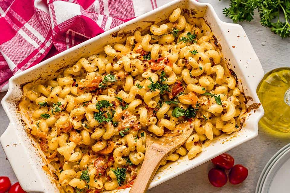 Baked pasta in a creamy sauce, topped with cherry tomatoes and fresh herbs, presented in a white dish, with a wooden spoon and a red-striped towel beside it.