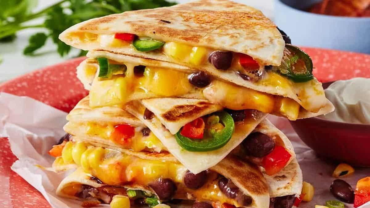 A stack of quesadilla wedges filled with melted cheese, black beans, corn, red pepper, and sliced jalapeños on a red plate with a side of sour cream.