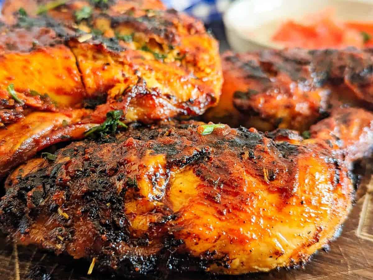Grilled chicken pieces with visible char marks, seasoned and garnished with herbs; a blurred bowl of sauce in the background.