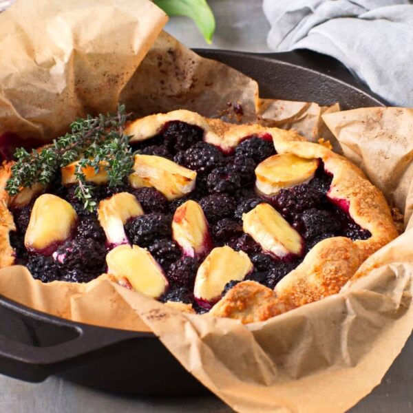 A rustic galette filled with blackberries and peach slices, garnished with sprigs of thyme, sits on parchment paper in a cast iron pan.