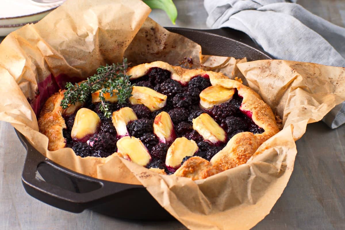 A rustic galette filled with blackberries and peach slices, garnished with sprigs of thyme, sits on parchment paper in a cast iron pan.