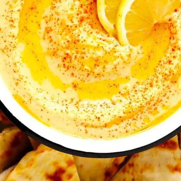 A bowl of hummus topped with olive oil and paprika, garnished with lemon slices, and surrounded by pieces of pita bread.
