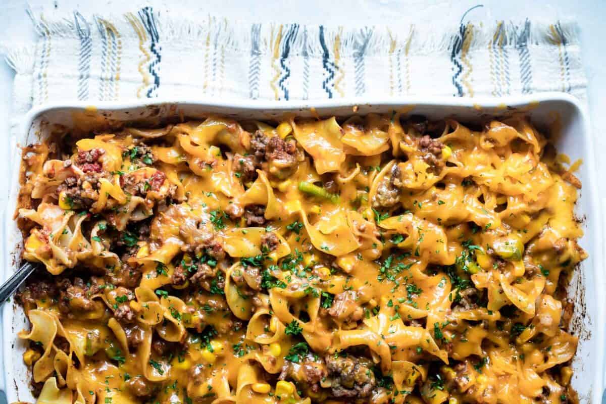 A baked casserole dish containing egg noodles, ground beef, and cheese, topped with melted cheddar cheese and chopped parsley.
