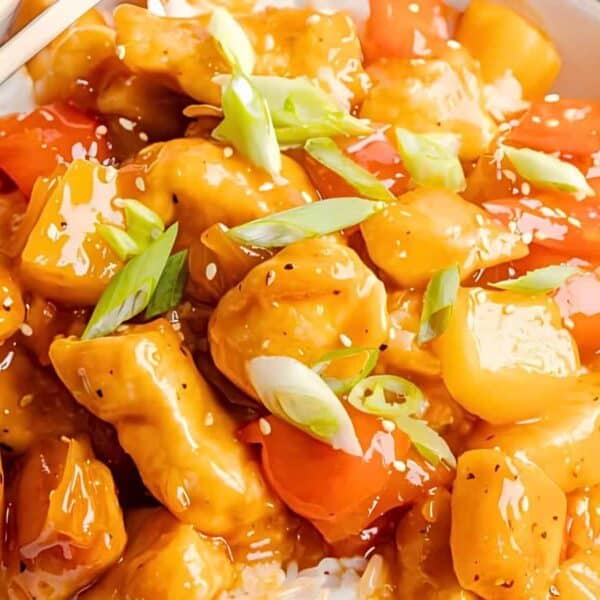 A bowl filled with pieces of chicken, red and yellow bell peppers, and rice, topped with sliced green onions. Chopsticks rest on the edge of the bowl.