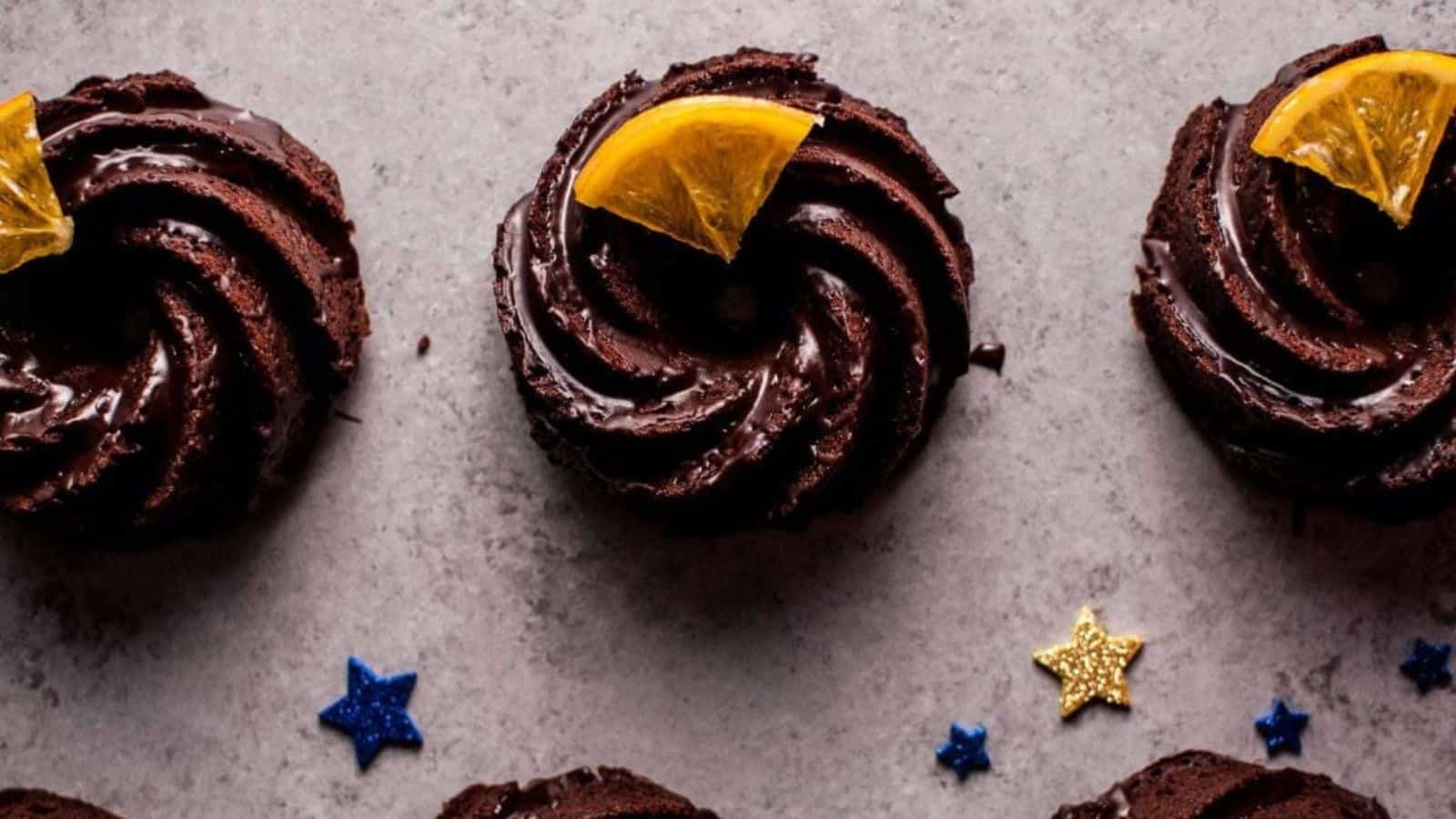 Chocolate mini bundt cakes topped with an orange slice are arranged in a row on a gray surface, adorned with blue and gold star-shaped decorations.