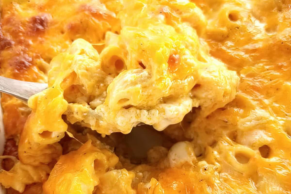 A close-up view of a creamy, cheesy macaroni and cheese dish being scooped with a spoon. The macaroni is covered in melted cheese, showcasing a golden, bubbly, and slightly crispy texture on top.