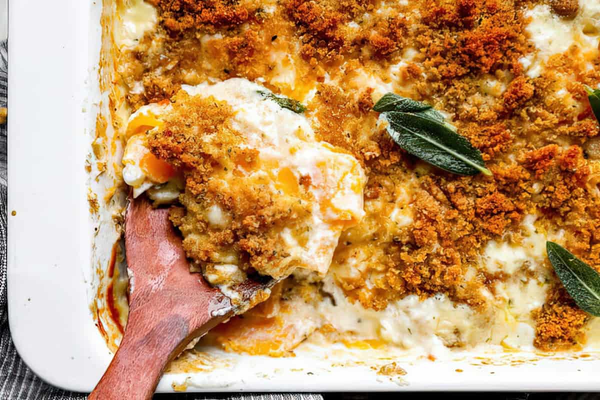 Close-up of a baked casserole dish with a wooden spoon scooping out a serving. The dish contains layers of creamy sauce, melted cheese, and a golden-brown breadcrumb topping with fresh sage leaves garnishing the surface.