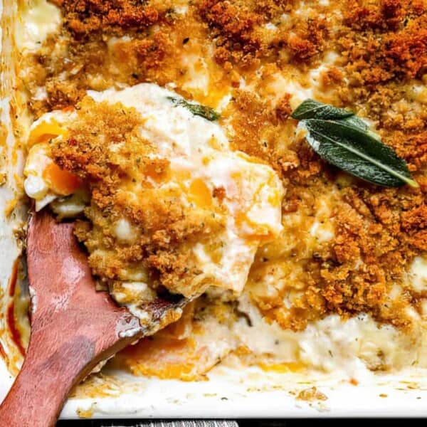 Close-up of a baked casserole dish with a wooden spoon scooping out a serving. The dish contains layers of creamy sauce, melted cheese, and a golden-brown breadcrumb topping with fresh sage leaves garnishing the surface.