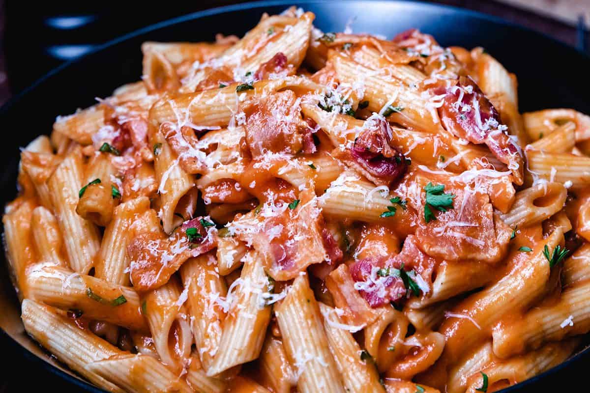 Penne pasta in a tomato-based sauce, garnished with grated cheese, bacon pieces, and fresh herbs in a black bowl.