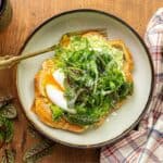 A plate of eggs on toast with avocado, garnished with greens, sits on a wooden table. Nearby are a plaid napkin, a pepper grinder, and a glass of red wine.