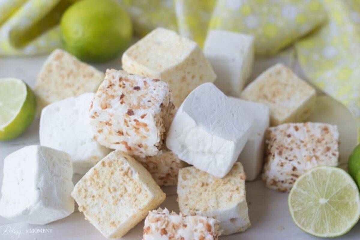 A stack of square marshmallows, some plain and some coated with toasted coconut, are displayed on a light surface. Fresh lime halves and a yellow patterned fabric are visible in the background, adding a touch of color to the arrangement.