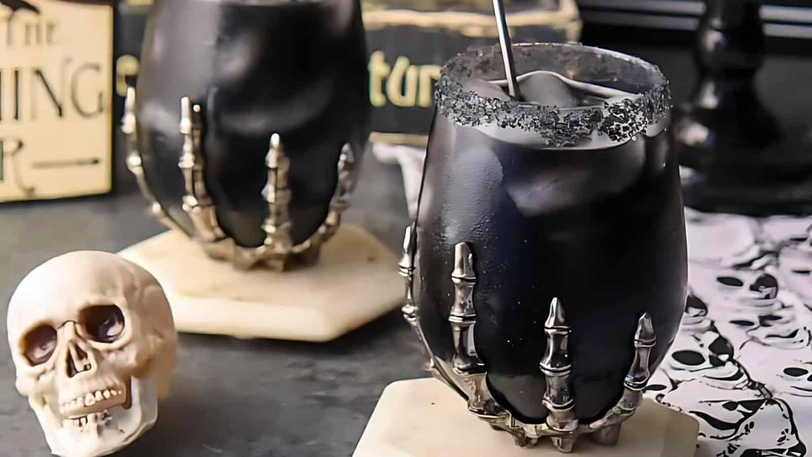 Two dark-colored drinks are served in glasses adorned with skeletal hand holders and rims coated in black sugar. The glasses are placed on white coasters, with one coaster also holding a small skull decoration. The background includes Halloween-themed items.