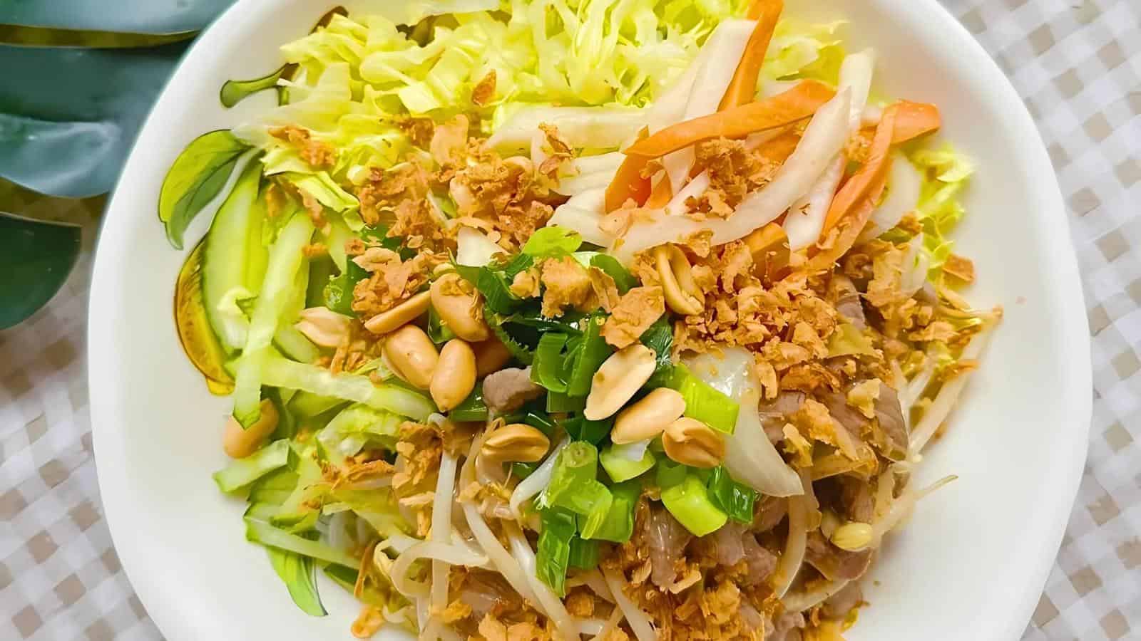 A plate of Vietnamese chicken salad featuring shredded lettuce, julienned carrots, cucumbers, bean sprouts, and chicken strips, topped with crushed peanuts, fried shallots, and herbs. The salad is arranged in a colorful and visually appealing manner.