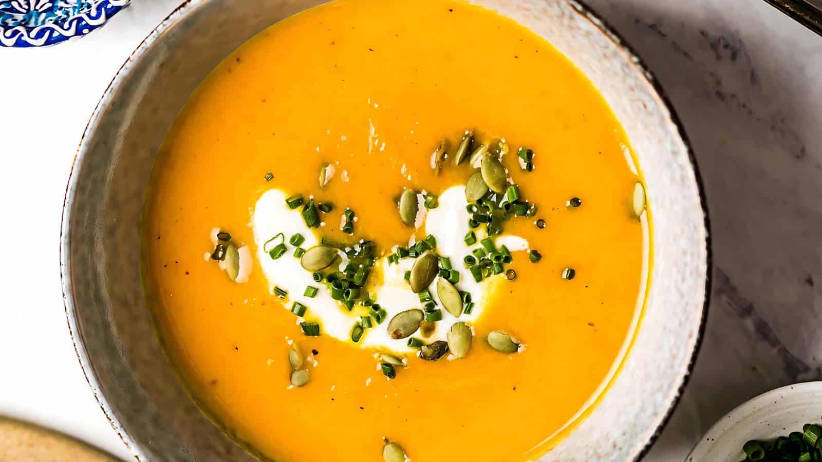 A bowl of creamy orange soup, garnished with a swirl of cream, chopped chives, pumpkin seeds, and black pepper. The soup appears smooth and is served in a simple, round bowl. The background features a partial view of a plate with a decorative pattern.