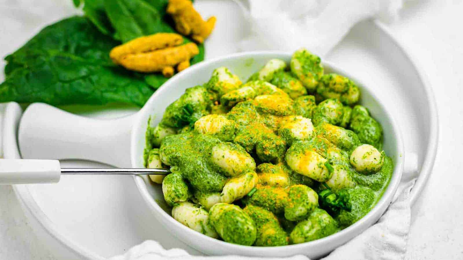 A white bowl filled with gnocchi covered in a green sauce, garnished with a sprinkle of orange seasoning. A fork rests on the side of the bowl. In the background, large spinach leaves and a couple of yellow flowers are visible on the white surface.