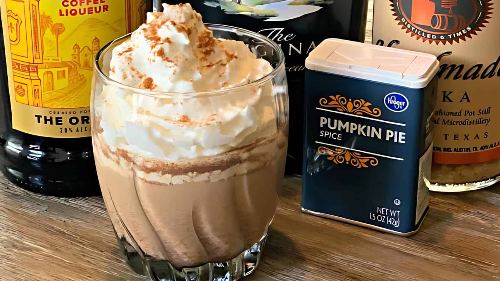 A glass filled with a creamy coffee drink topped with a generous amount of whipped cream and sprinkled with pumpkin pie spice, surrounded by bottles of coffee liqueur and vodka, as well as a tin of pumpkin pie spice on a wooden surface.