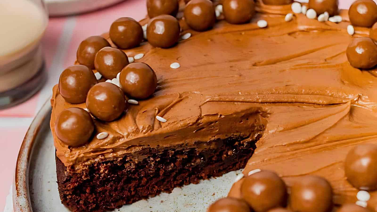 A close-up of a chocolate cake with a smooth frosting, topped with round chocolate candies and a few white sprinkles. A slice has been cut out, revealing a moist and rich interior. The cake sits on a white plate with a blurred background.