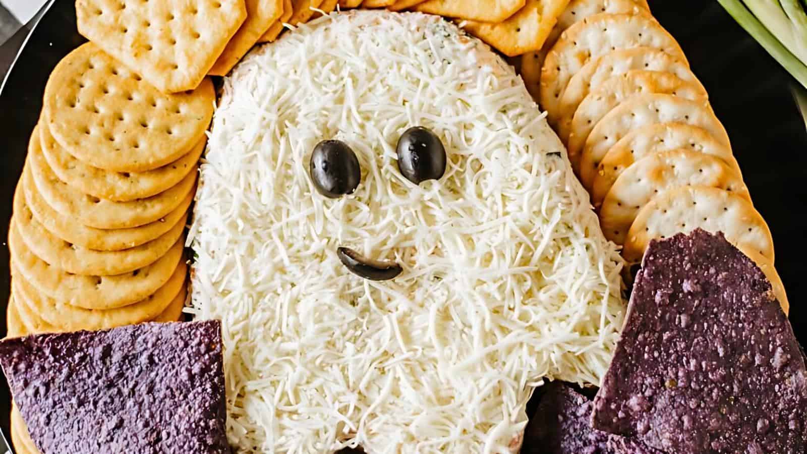 A Halloween-themed snack platter features a ghost-shaped dip covered in shredded cheese, with black olives for eyes and a smile made of a small piece of olive. The ghost is surrounded by round crackers and triangular purple tortilla chips.