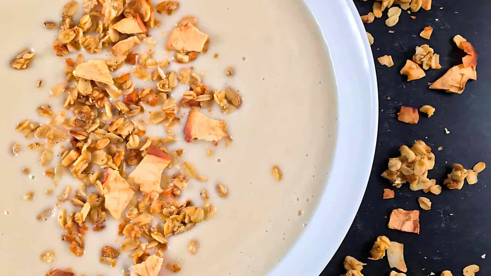 A white bowl filled with creamy beige soup garnished with granola and small chunks of dried fruit. Some granola and fruit pieces are scattered on the dark surface next to the bowl.