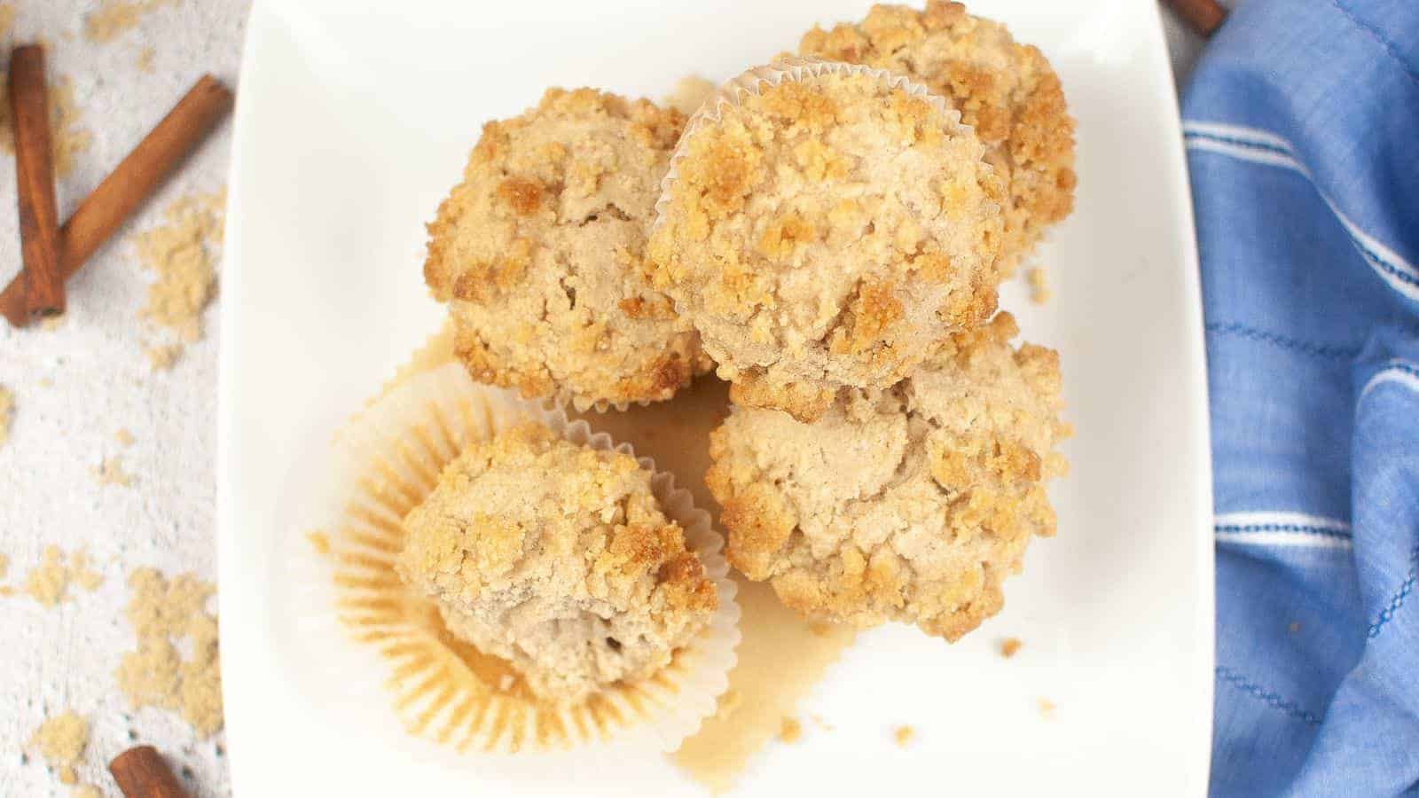 A white square plate holds five crumb-topped muffins, some in paper liners. The plate is placed on a light surface, with two cinnamon sticks nearby and a blue and white cloth to the right. Crumbs are scattered around, hinting at a soft and crumbly texture.