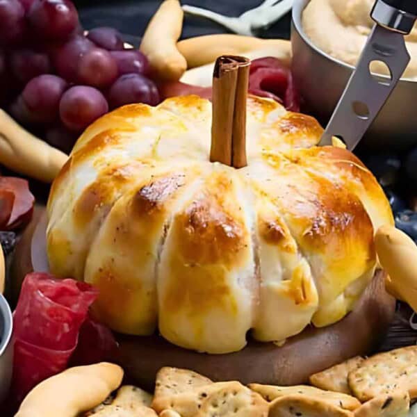 A platter featuring a pumpkin-shaped baked brie in the center, surrounded by various foods including grapes, crackers, meats, cheeses, breadsticks, and a bowl of red jam. A small knife is inserted into the baked brie, highlighting its centerpiece role.