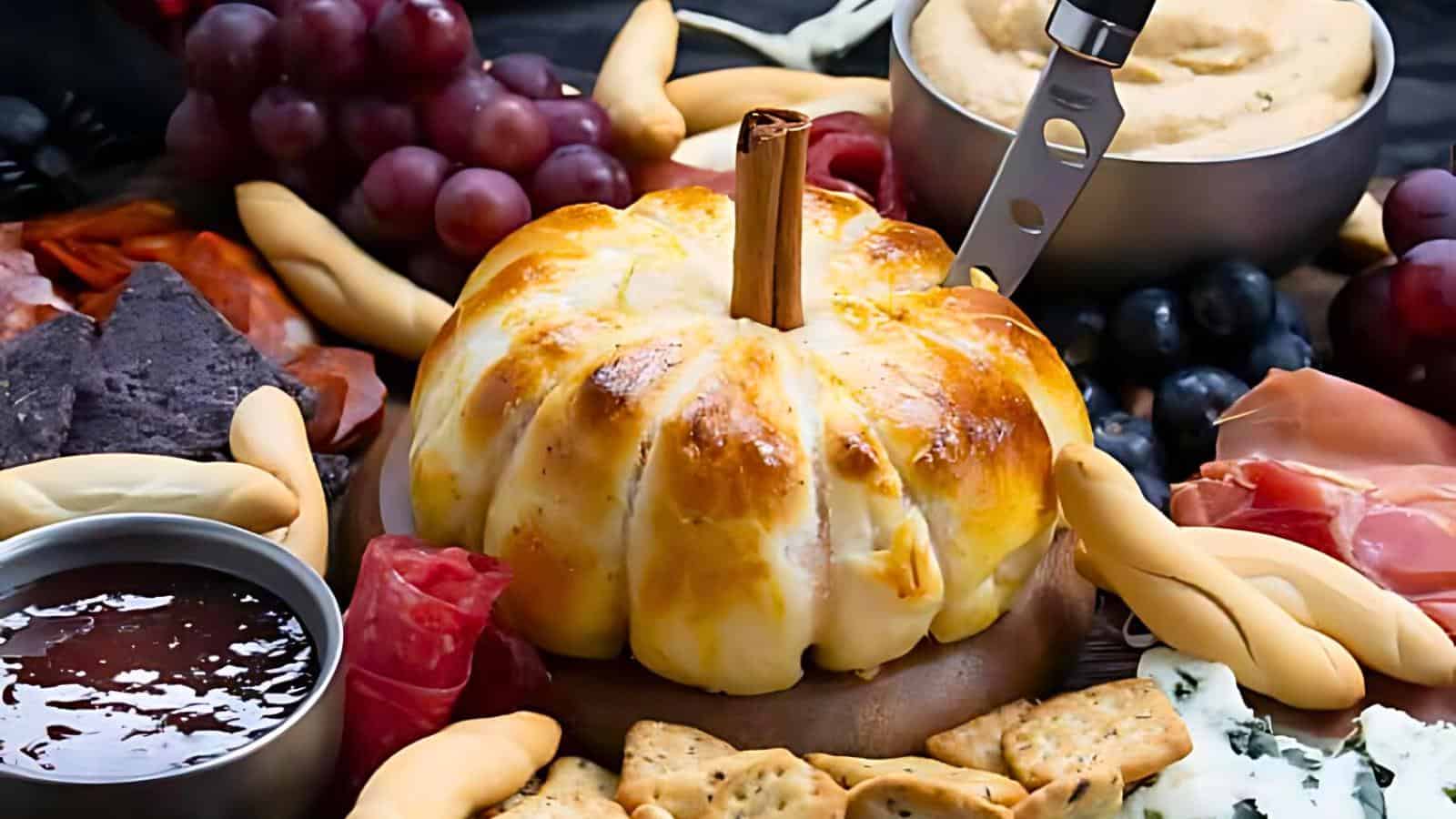 A platter featuring a pumpkin-shaped baked brie in the center, surrounded by various foods including grapes, crackers, meats, cheeses, breadsticks, and a bowl of red jam. A small knife is inserted into the baked brie, highlighting its centerpiece role.