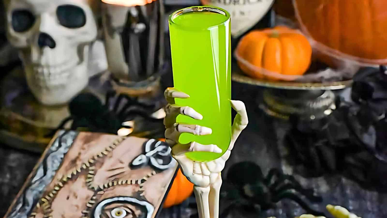 A tall, glowing green drink is held by a skeletal hand prop. The background features Halloween-themed decorations including a skull, pumpkins, spiders, and a spellbook with a stitched face cover. The scene is dimly lit, enhancing the spooky ambiance.