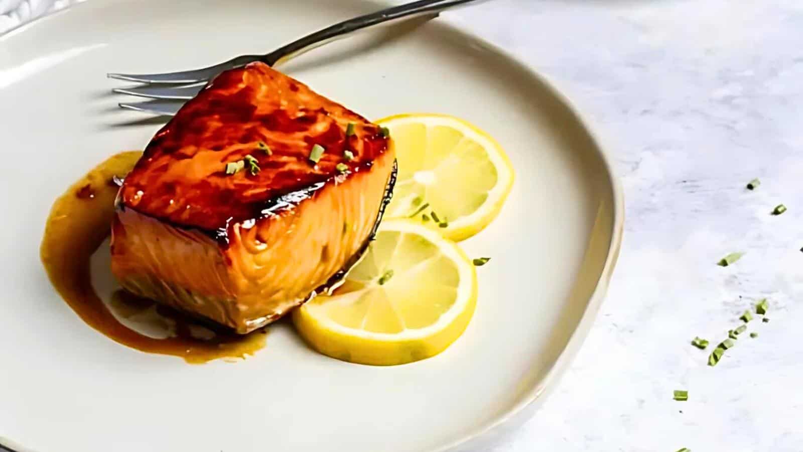 A piece of glazed salmon, garnished with finely chopped herbs, sits on a light-colored plate next to two slices of lemon. A fork rests at the top of the plate. The background is a white, marbled surface.