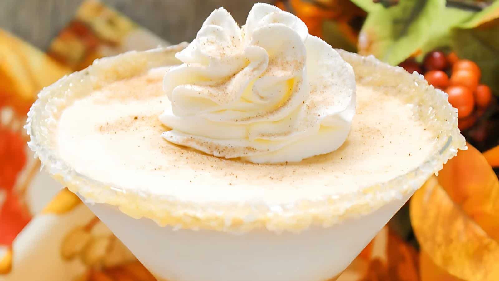 A close-up shot of a creamy cocktail in a martini glass with a sugar-rimmed edge, topped with a swirl of whipped cream and a sprinkle of cinnamon. Autumn leaves and berries are blurred in the background.