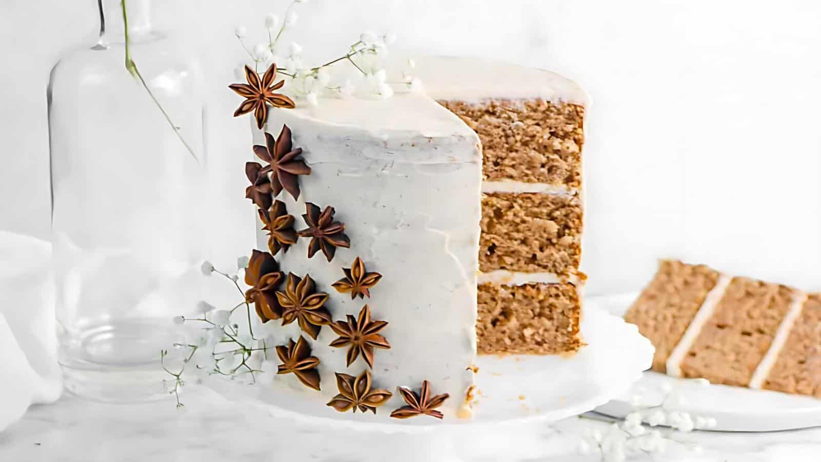 A white frosted, three-layer cake decorated with star anise and delicate white flowers is displayed on a cake stand. A slice of the cake is placed beside it on a plate, showing the light brown interior. The background is a minimalistic white setting.