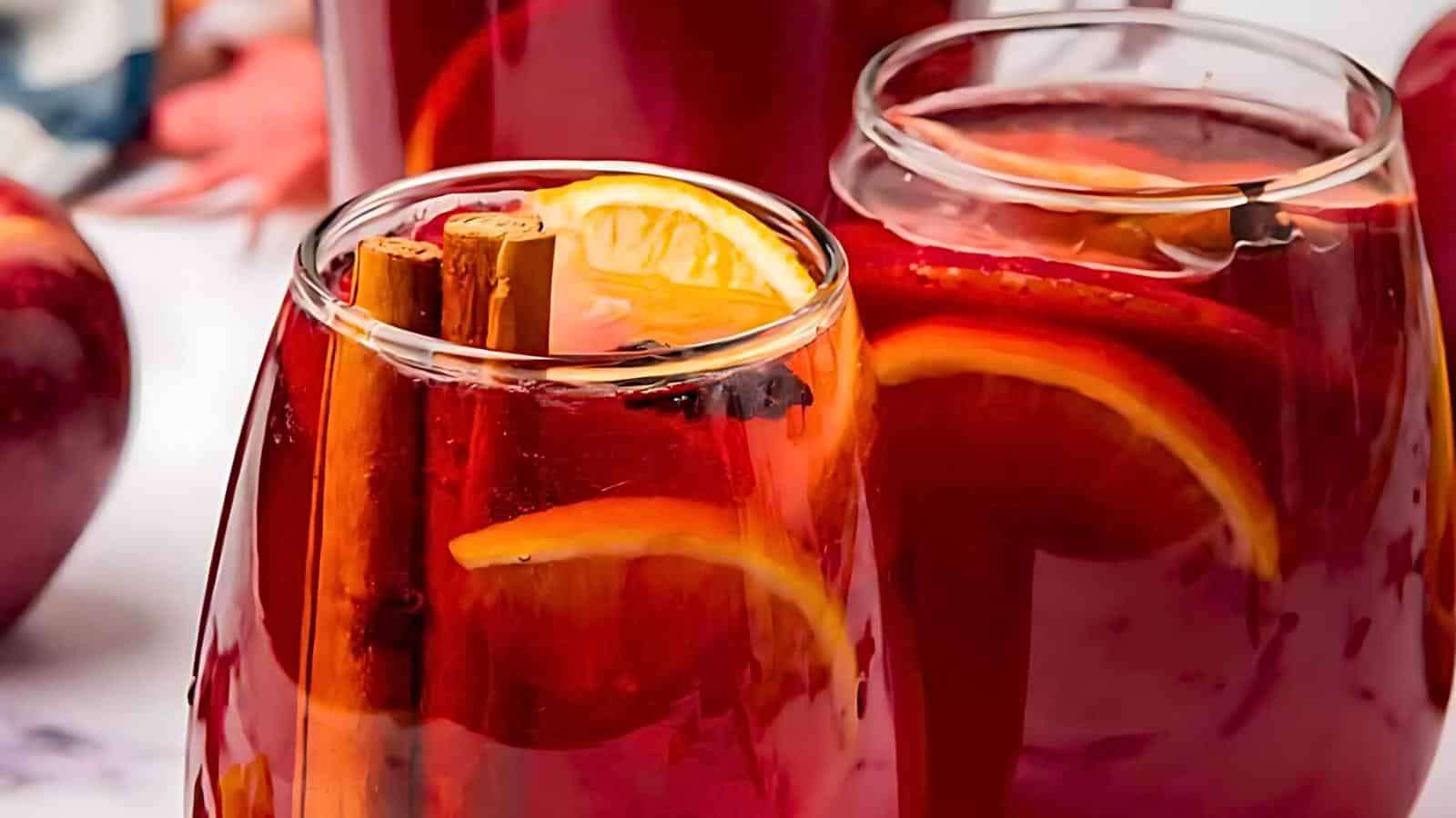 Close-up of three glasses filled with red sangria. Each glass contains slices of orange and sticks of cinnamon, with a rich reddish hue. The background features red apples and another glass of sangria, providing a vibrant, refreshing, and festive visual.