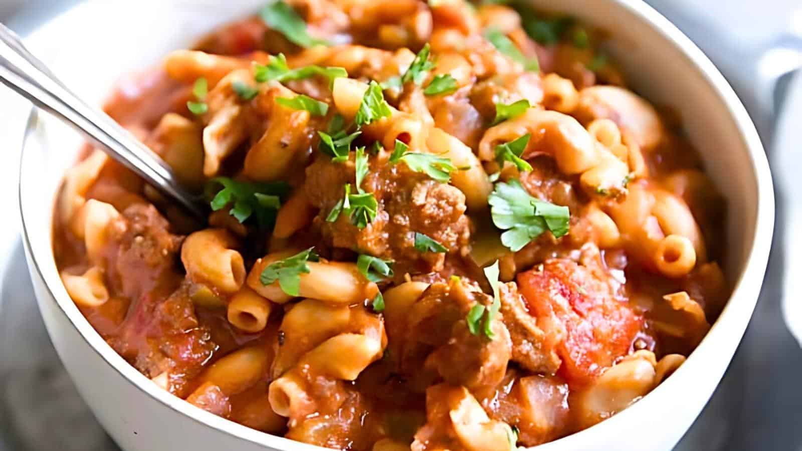 A bowl of goulash pasta featuring a rich, chunky tomato sauce with ground meat and macaroni noodles, garnished with fresh parsley. A spoon is partially submerged in the hearty dish, ready for a bite.