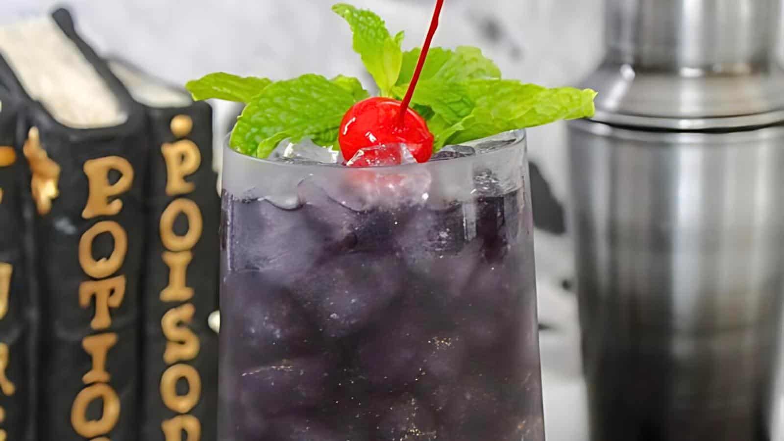 A tall glass filled with a dark purple iced beverage, topped with a red cherry and a sprig of mint. Behind the glass, there are two stacked books with visible text on the spine and a metal cocktail shaker. The background is out of focus.