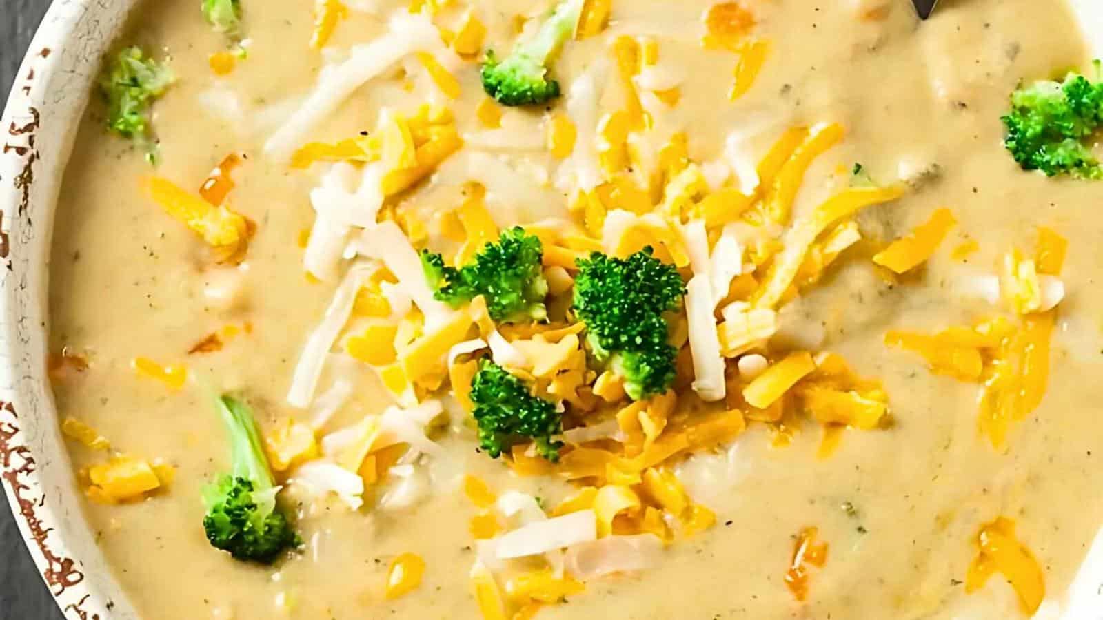A top-down view of a bowl of creamy broccoli cheddar soup. The thick soup is garnished with shredded cheddar cheese, white cheese, and small broccoli florets. The soup has a smooth, rich texture with visible pieces of cheese and broccoli.