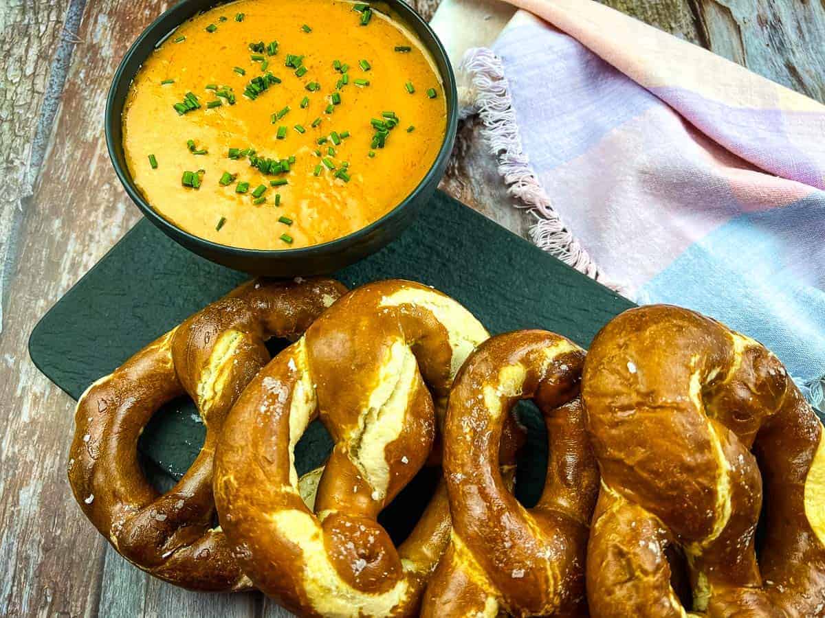 A tray of freshly baked soft pretzels is displayed next to a bowl of creamy cheddar cheese sauce garnished with chopped chives. A colorful cloth is partially visible in the background on a rustic wooden surface.