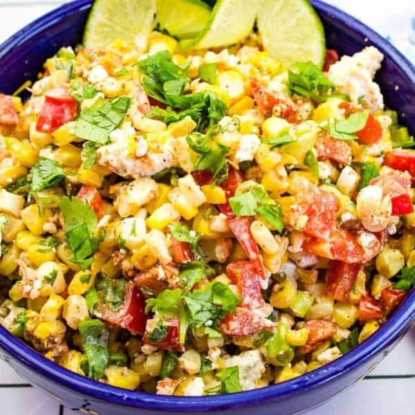 A vibrant bowl of Mexican street corn salad (Esquites) filled with roasted corn, diced red bell peppers, crumbled cheese, and fresh cilantro. Lime wedges garnish the top, and the colorful ingredients are mixed in a deep blue bowl.