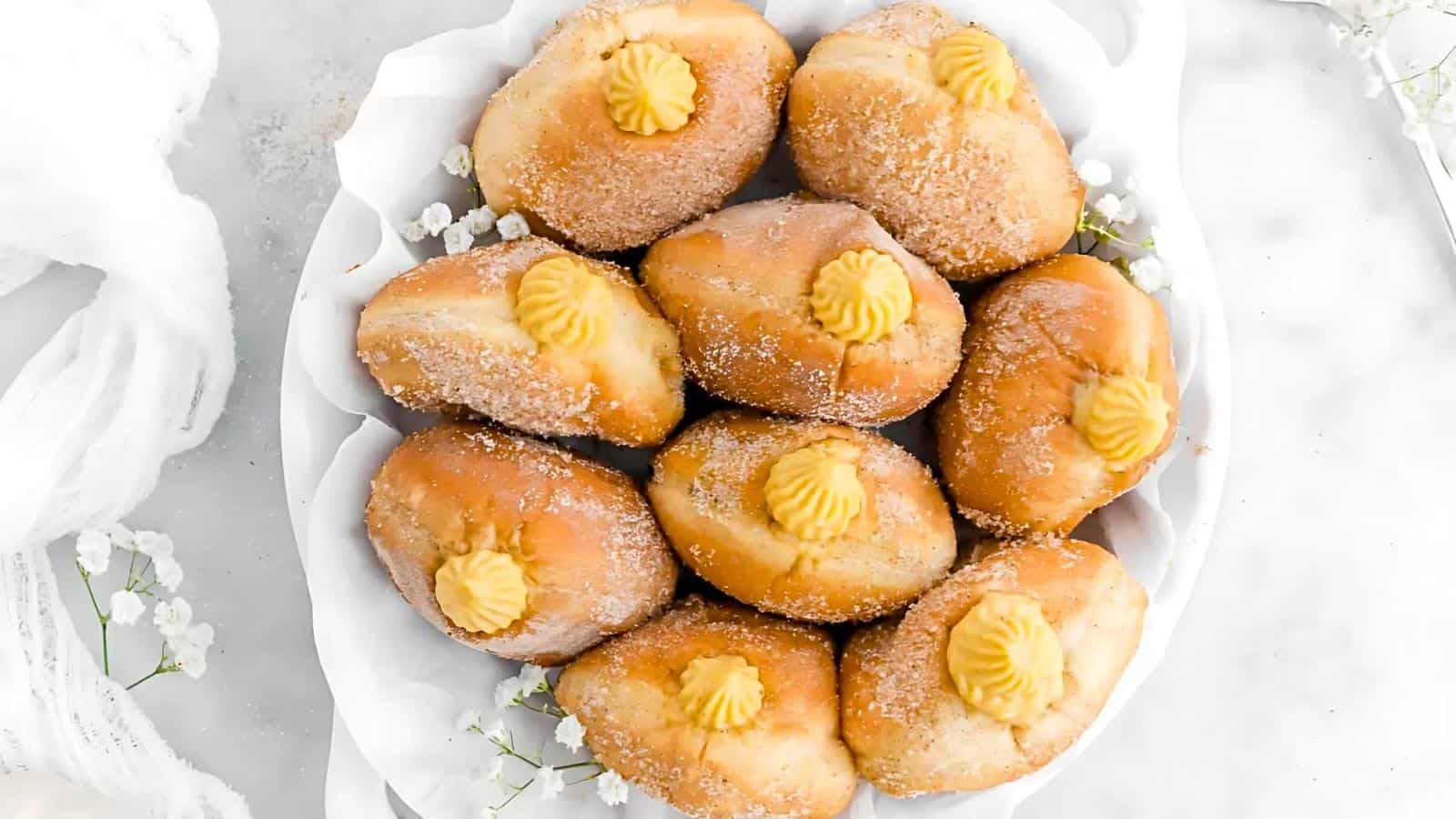 A white plate of sugar-coated doughnuts filled with a creamy yellow custard is arranged on a light-colored surface. The doughnuts are garnished with white flowers for decoration.