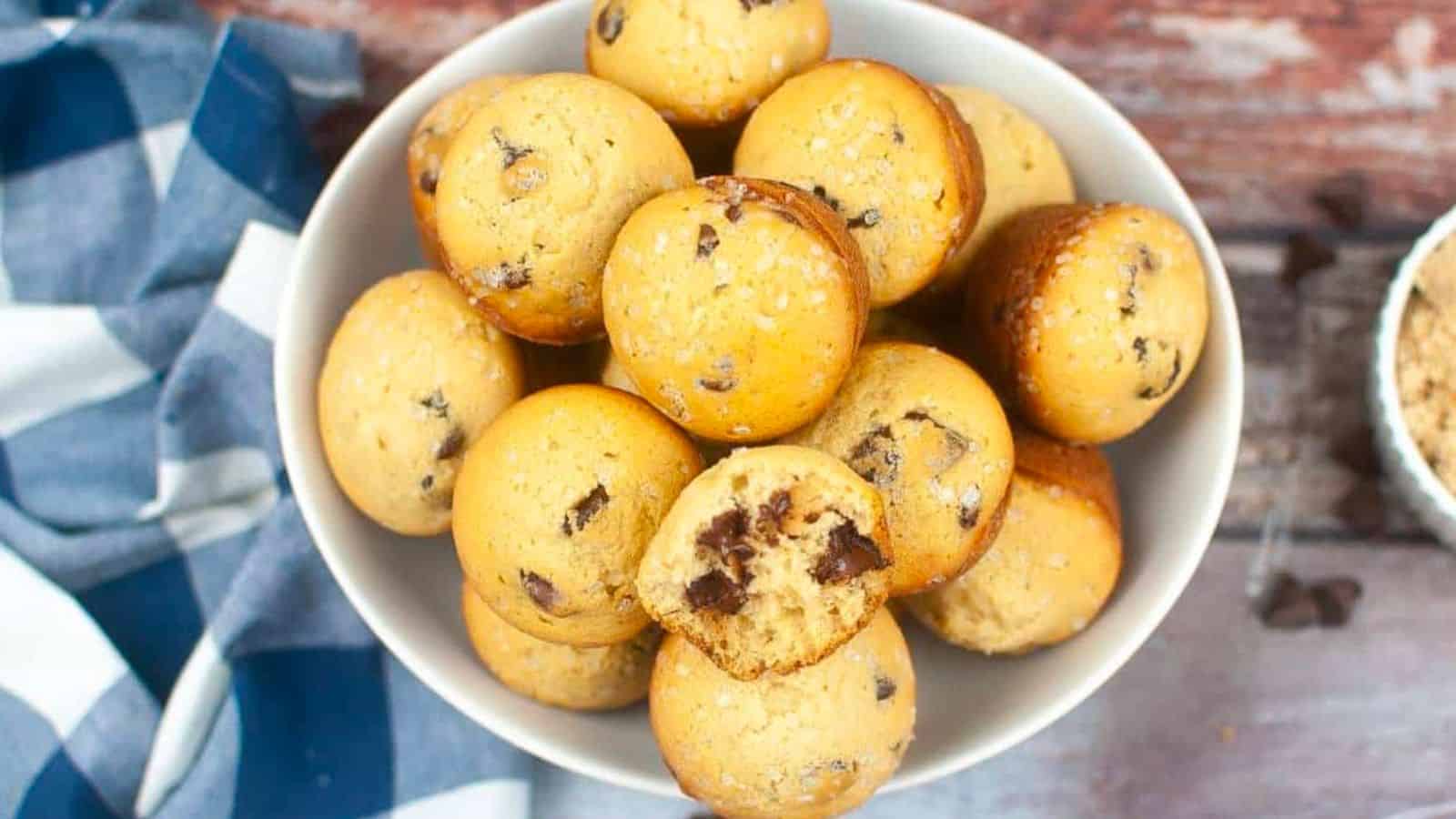 A bowl filled with golden-brown chocolate chip muffins, with one muffin showing a bite taken out, revealing melted chocolate chips inside. The bowl is placed on a rustic wooden surface with a blue and white checkered cloth and a small bowl of crushed ingredients nearby.