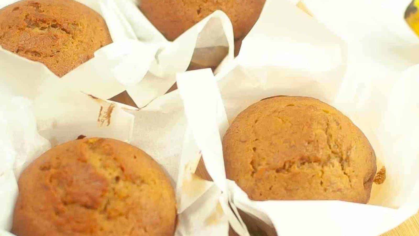 Four freshly baked muffins are wrapped in parchment paper. They are golden brown with slightly cracked tops, indicating a moist and fluffy texture inside. The muffins are arranged closely together, highlighting their homemade and inviting appearance.