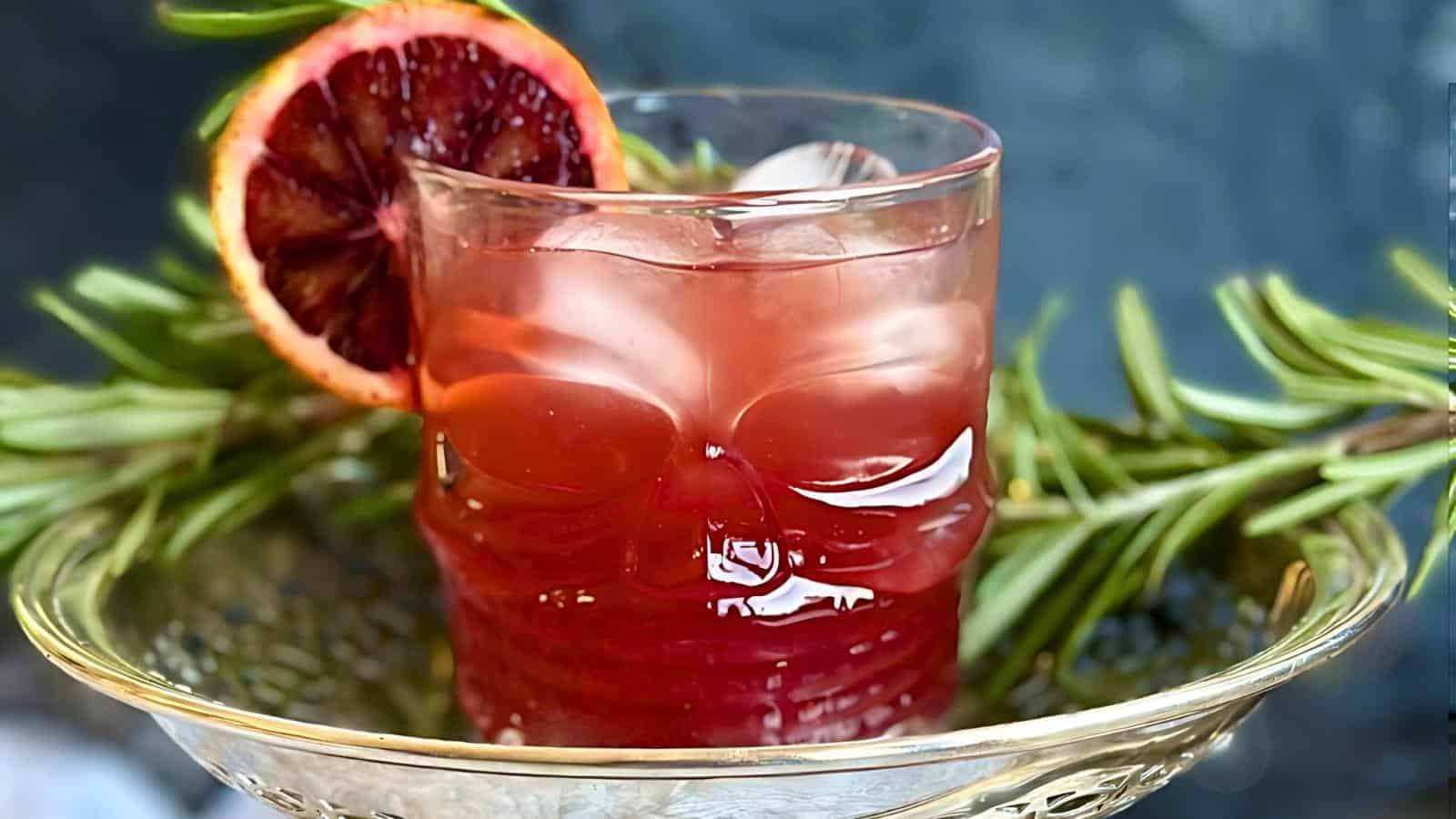 A dark red cocktail in a skull-shaped glass, filled with ice and garnished with a slice of blood orange. The glass is placed on a decorative clear plate, surrounded by sprigs of rosemary. The background is dark and blurred.